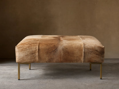 Tilton Hair on Hide Ottoman, rectangular, upholstered in blonde brindle hair-on-hide with gold metal legs, on wood floor.
