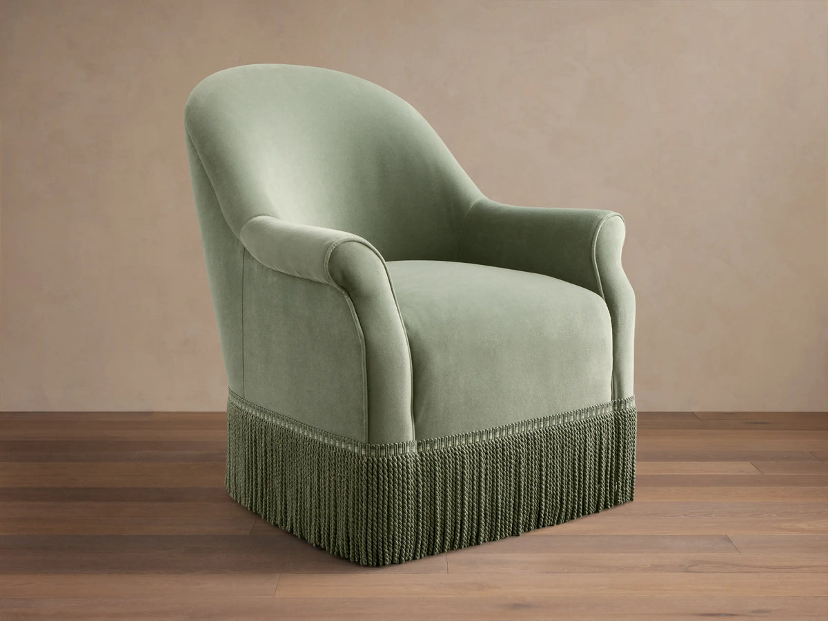 Front view of the sage green Augustus Swivel Chair with Fringe, featuring a rounded back, rolled arms, and a thick green fringe base.