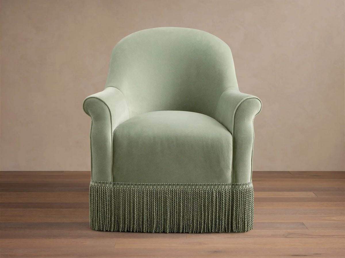 Front view of the sage green Augustus Swivel Chair with Fringe, featuring a rounded back, rolled arms, and a thick green fringe base.