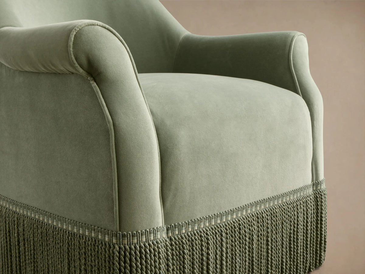 Rear-side view of the sage green Augustus Swivel Chair with Fringe, with a rolled arm and full fringe detail at the base.