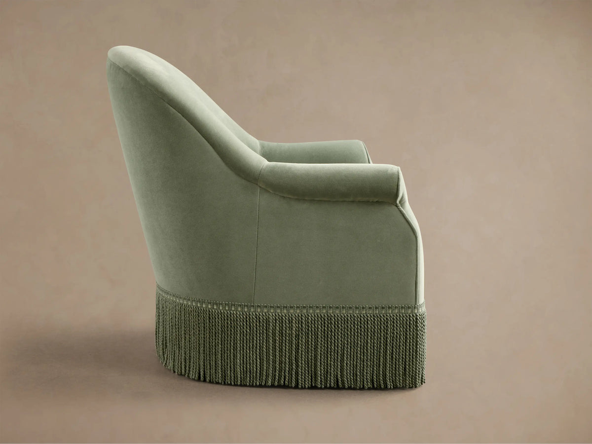 Close-up, angled view of a sage green Augustus Swivel Chair with Fringe, highlighting the rolled armrest and braided fringe.
