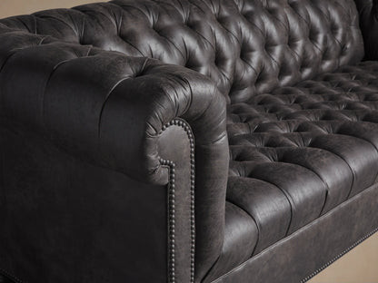 Berwick Faux Leather Sofa, Norre Steel, with deep tufting, rolled arms, nailhead trim, and turned dark legs on a wood floor.