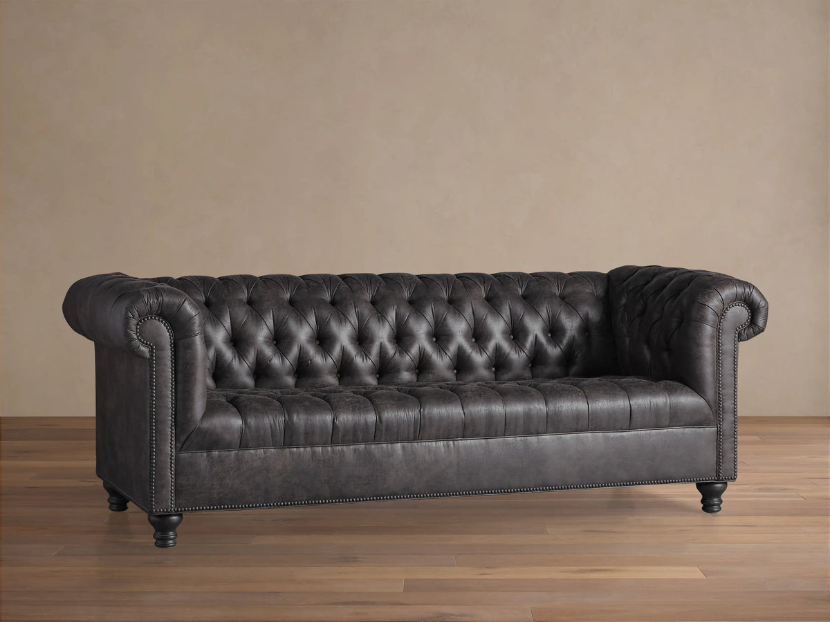 Berwick Faux Leather Sofa in dark gray with deep button tufting, rolled arms, nailhead trim, and dark wooden legs.