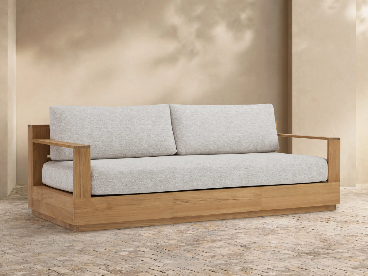 Bal Harbour outdoor teak sofa with light grey cushions, centered against a textured tan wall and patterned stone floor.
