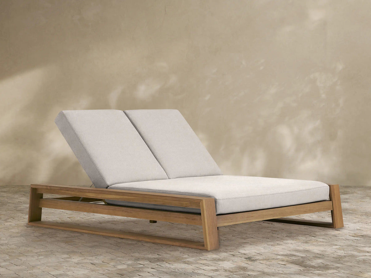 Canyon Outdoor Teak Double Chaise with natural wood frame and light beige cushions, both backrests inclined, angled view on white.