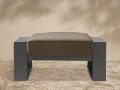 Canyon Outdoor Aluminum Ottoman: Dark gray aluminum frame, light brown cushion with rounded edges, front view on white background.