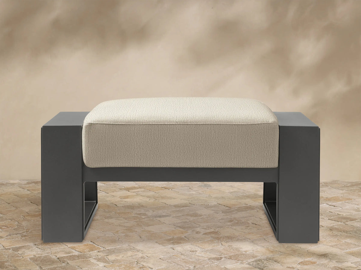 Canyon Outdoor Aluminum Ottoman, front view. Dark grey rectangular aluminum frame with a thick, light beige cushion with rounded corners.