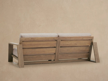Front view of the Canyon Outdoor Teak Sofa with natural teak frame and light beige cushions on a paved patio.