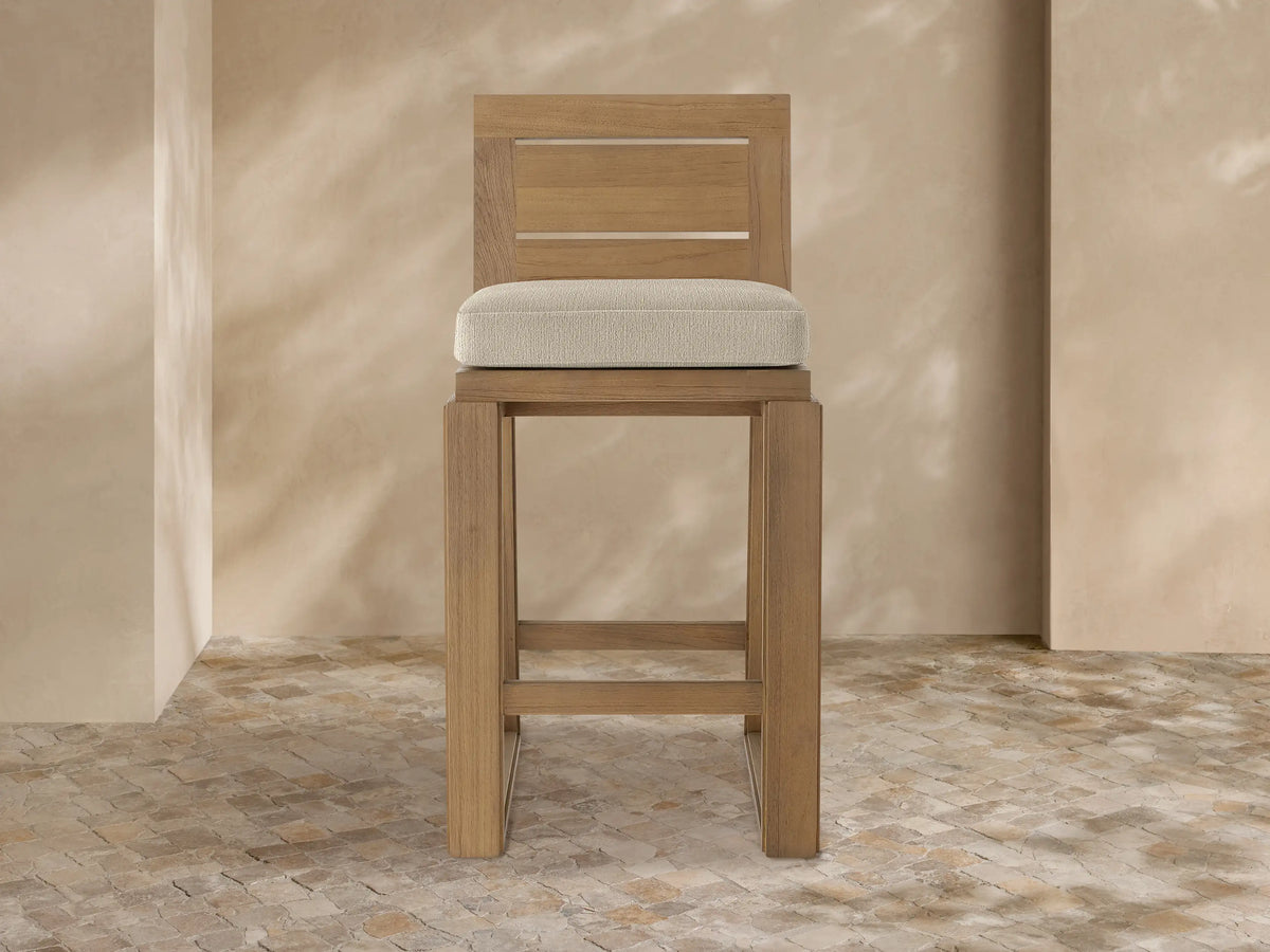 Canyon Outdoor Teak Barstool, front view. Light brown teak wood frame, slatted back, and a beige upholstered seat cushion.