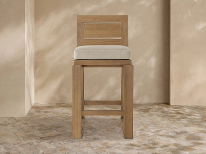 Canyon Outdoor Teak Barstool, front view. Light brown teak wood frame, slatted back, and a beige upholstered seat cushion.