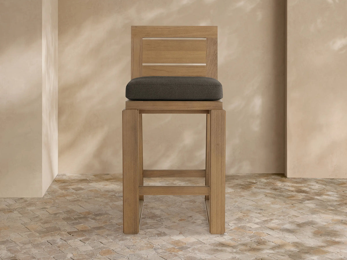 Front view of Canyon Outdoor Teak Barstool, light brown teak frame with two back slats and dark gray seat cushion.