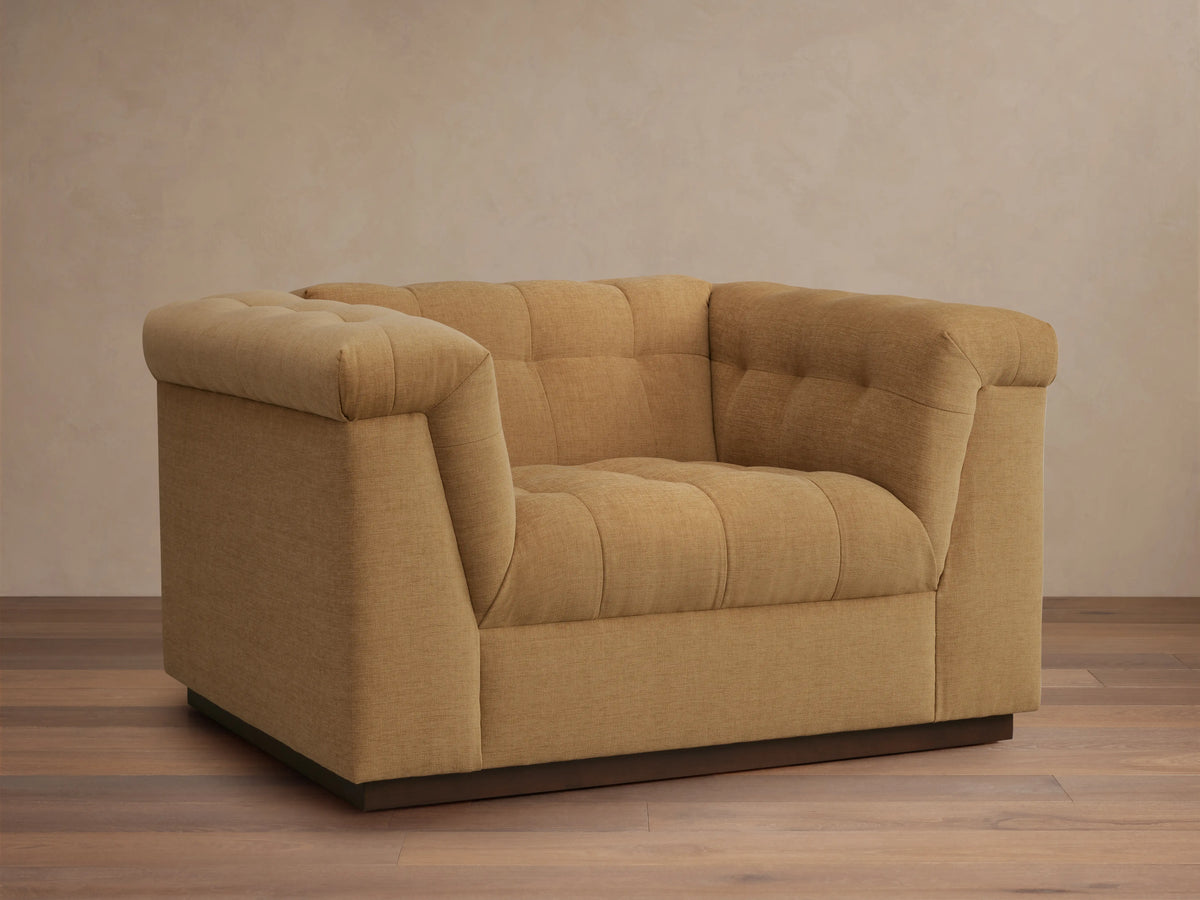 Cameran Chair in light brown fabric, featuring tufted backrests and cushioned armrests, set on a wooden floor against a beige wall.