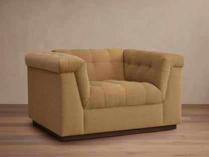 Cameran Chair in light brown fabric, featuring tufted backrests and cushioned armrests, set on a wooden floor against a beige wall.