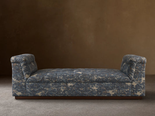 Daybed with patterned upholstery in blue and gray tones, featuring cushioned backrests on each end and set against a neutral wall.