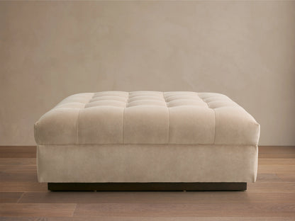 Cameran Ottoman in beige fabric with tufted top, rectangular shape, on a wooden floor, against a neutral wall.