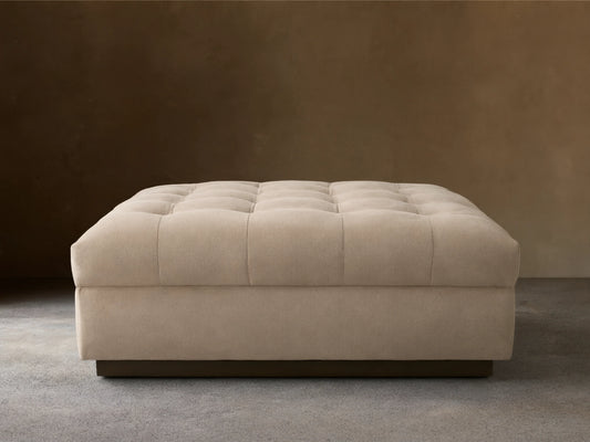 Cameran Ottoman in beige fabric with tufted top, rectangular shape, on a wooden floor, against a neutral wall.