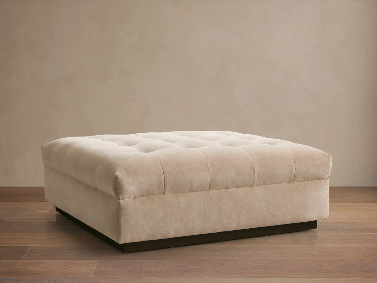 Cameran Ottoman in beige fabric with tufted top, rectangular shape, on a wooden floor, against a neutral wall.