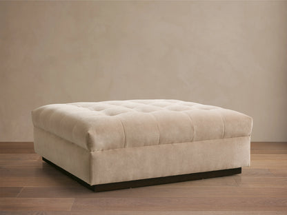 Cameran Ottoman in beige fabric with tufted top, rectangular shape, on a wooden floor, against a neutral wall.