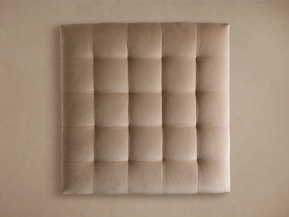 The Cameran Ottoman features a light beige, tufted top with soft fabric, and a rectangular shape on a dark wood base, set on wooden flooring.