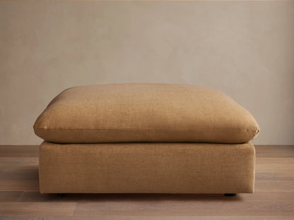Calistoga Ottoman in light brown fabric with a plush cushion top, resting on a wooden floor, against a beige wall.