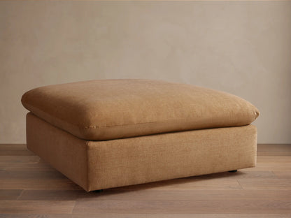 Calistoga Ottoman in light brown fabric with a plush cushion top, resting on a wooden floor, against a beige wall.