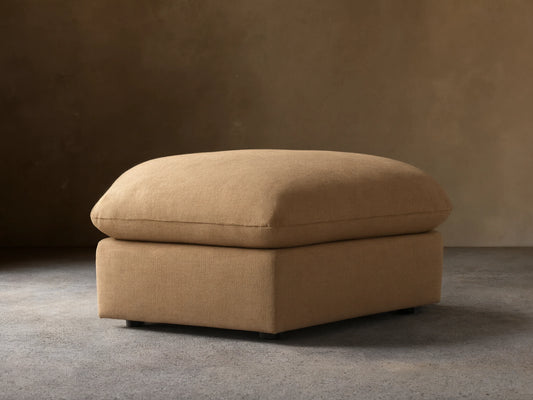 Calistoga Ottoman in light brown fabric with a plush cushion top, resting on a wooden floor, against a beige wall.