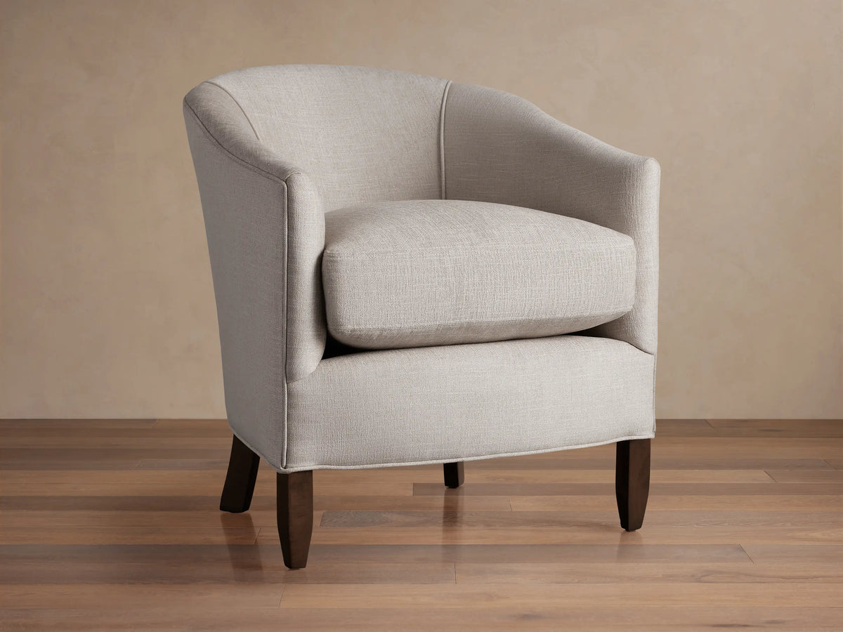 Giles Chair: Light neutral fabric armchair, curved back, separate seat cushion, dark wood legs. Angled slightly right on wood floor.