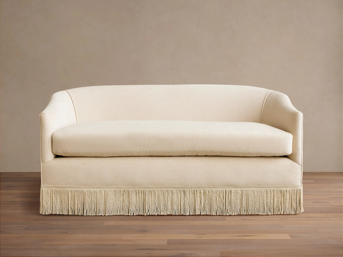 Cream-colored Giles Settee with rounded arms, a single seat cushion, and a fringe skirt, set on a wooden floor.