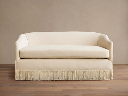 Cream-colored Giles Settee with rounded arms, a single seat cushion, and a fringe skirt, set on a wooden floor.