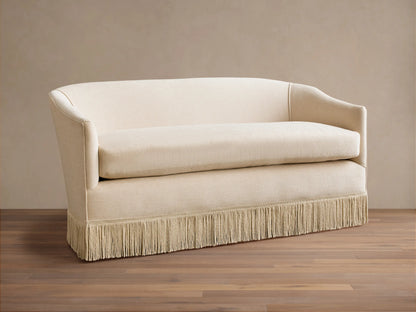 Cream-colored Giles Settee with rounded arms, a single seat cushion, and a fringe skirt, set on a wooden floor.
