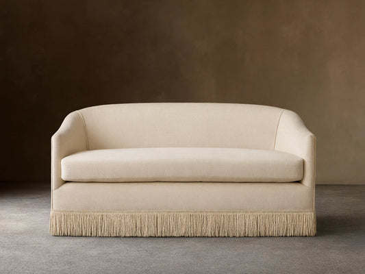 Cream-colored Giles Settee with rounded arms, a single seat cushion, and a fringe skirt, set on a wooden floor.