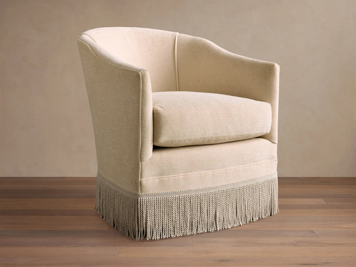 A beige Giles Swivel Chair with a curved back, plush cushion, and decorative fringe around the base, on a polished wood floor.