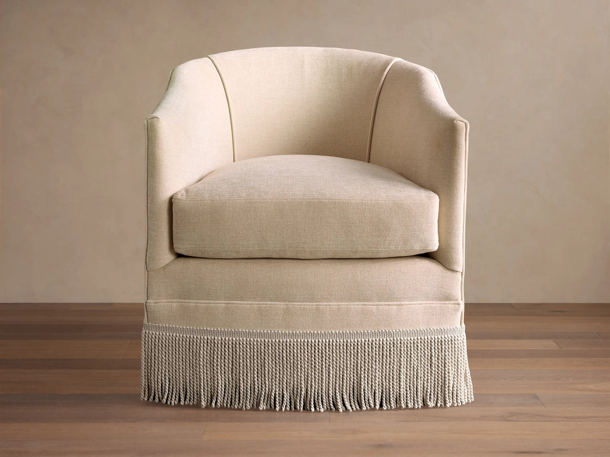 A beige Giles Swivel Chair with a curved back, plush cushion, and decorative fringe around the base, on a polished wood floor.