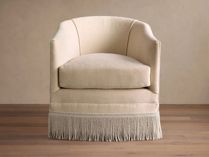 A beige Giles Swivel Chair with a curved back, plush cushion, and decorative fringe around the base, on a polished wood floor.