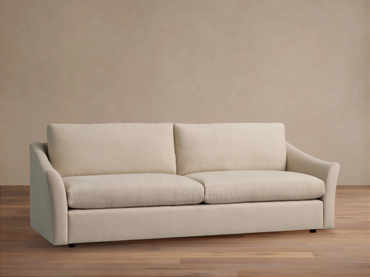 Helena Sofa, a light beige two-seater sofa with two back and seat cushions, flared arms, against a beige wall on a wooden floor.