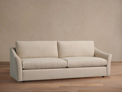 Helena Sofa, a light beige two-seater sofa with two back and seat cushions, flared arms, against a beige wall on a wooden floor.