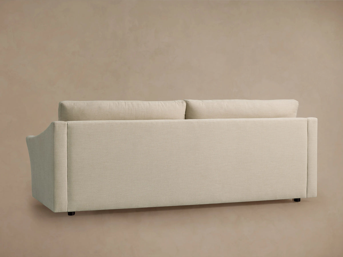 Back view of the Helena Sofa, upholstered in light cream fabric with two plush back cushions and small black feet, against a beige wall.