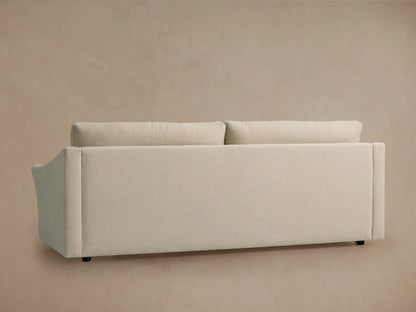 Back view of the Helena Sofa, upholstered in light cream fabric with two plush back cushions and small black feet, against a beige wall.