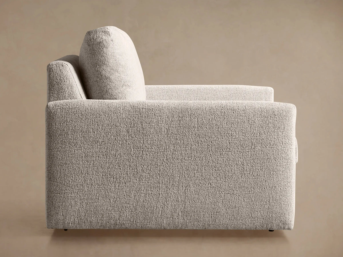 Kipton Slim Arm Chair upholstered in light grey textured fabric, seen from the back and side against a warm tan background.