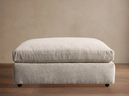 Light neutral textured Kipton Square Ottoman with a plush cushion top, resting on a wooden floor. Small dark round feet visible.
