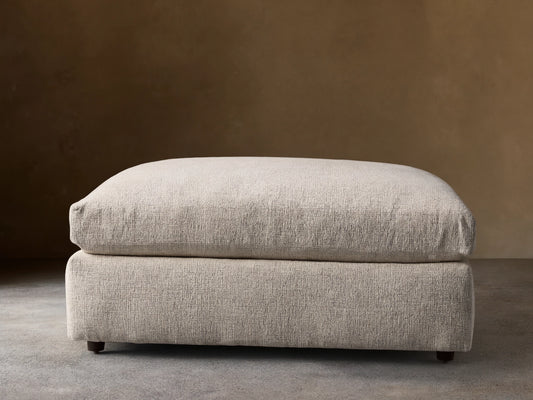 Light neutral textured Kipton Square Ottoman with a plush cushion top, resting on a wooden floor. Small dark round feet visible.