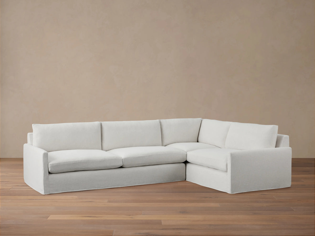 A light grey Kipton Slipcovered Two-Piece Sectional with a right-facing chaise. It has a soft, tailored appearance on a wooden floor.