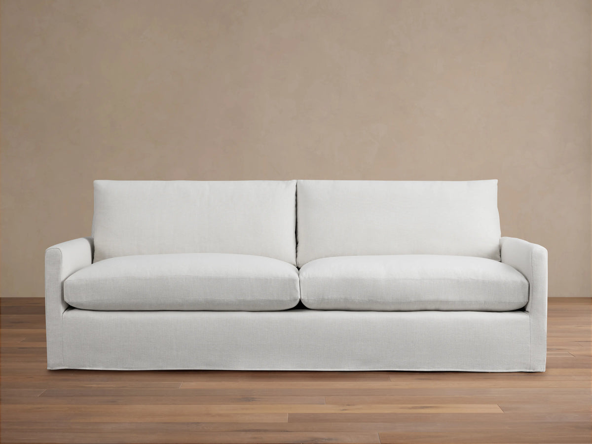Kipton Slipcovered Sofa in light beige fabric. Features straight arms, deep seat cushions, and a minimalist design on wooden floor.