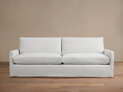 Kipton Slipcovered Sofa in light beige fabric. Features straight arms, deep seat cushions, and a minimalist design on wooden floor.