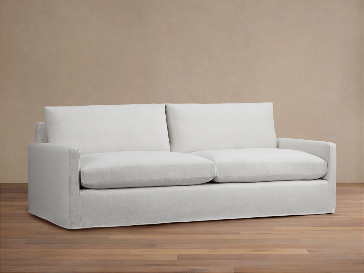 Kipton Slipcovered Sofa in light beige fabric. Features straight arms, deep seat cushions, and a minimalist design on wooden floor.