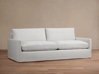 Kipton Slipcovered Sofa in light beige fabric. Features straight arms, deep seat cushions, and a minimalist design on wooden floor.