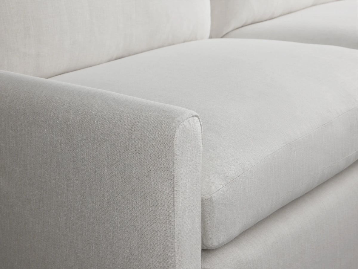A light gray Kipton Slipcovered Sofa on a wooden floor, featuring a sleek, modern design with two large seat cushions and backrests, in a minimalistic room.