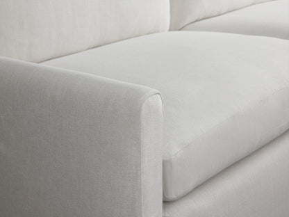 A light gray Kipton Slipcovered Sofa on a wooden floor, featuring a sleek, modern design with two large seat cushions and backrests, in a minimalistic room.