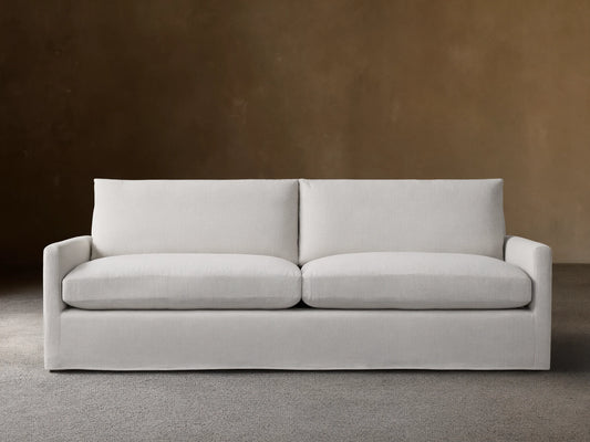 Kipton Slipcovered Sofa in light beige fabric. Features straight arms, deep seat cushions, and a minimalist design on wooden floor.
