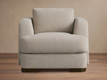 Beige upholstered Kayden Chair on a wooden floor, featuring a boxy design with thick, comfortable cushions and rounded armrests.